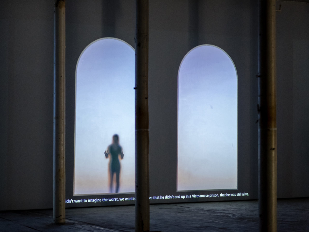A dark lit room with two projected arches on the wall, with a figure of someone standing holding their handing up against the flat surface. There is a passage of writing on the bottom of the page.