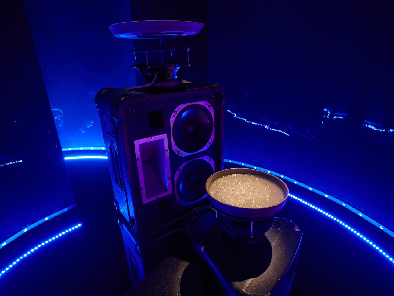 A sound installation with a speaker and a bowl of vibrating water, the space is lit up with blue lighting