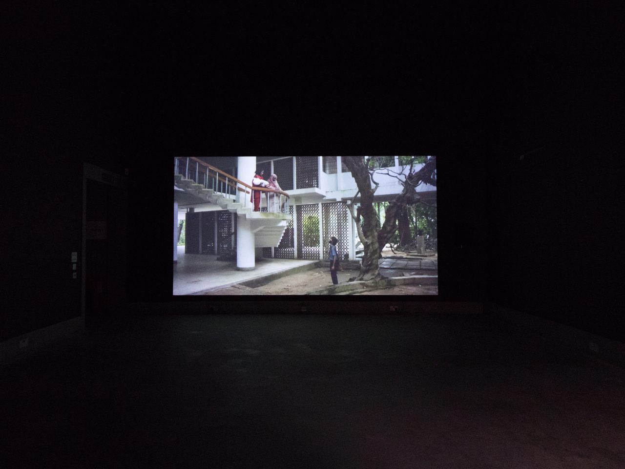 An installation shot of a film projected onto a wall. There are people looking down at someone from a staircase.