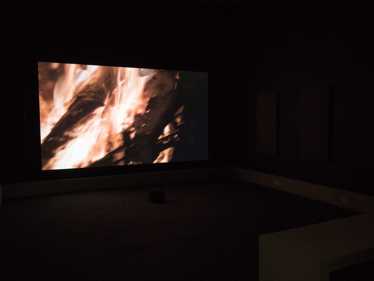 A installation shot of a film playing, the screen presents a close up shot of a bonfire.