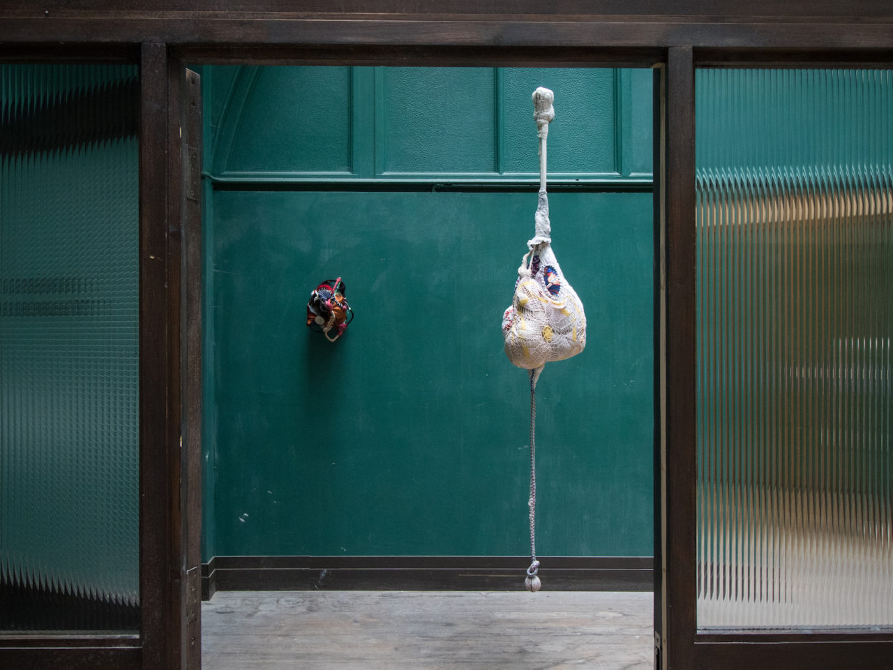 Two fabric sculptures, one on the wall and the other hanging. The wall sculpture is round and colourful and the hanging is long ans mainly white.
