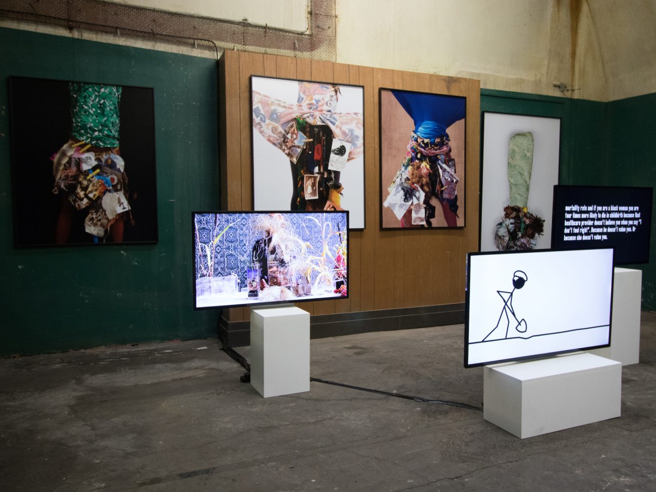 Four large flat fashion photographs on the walls of Cotton Exchange, the space had dark green walls with a wooden space in the middle. There are 3 screens in the foreground, one with a stick man animation, one with information text and the other of a woman with her hands in a bowl.