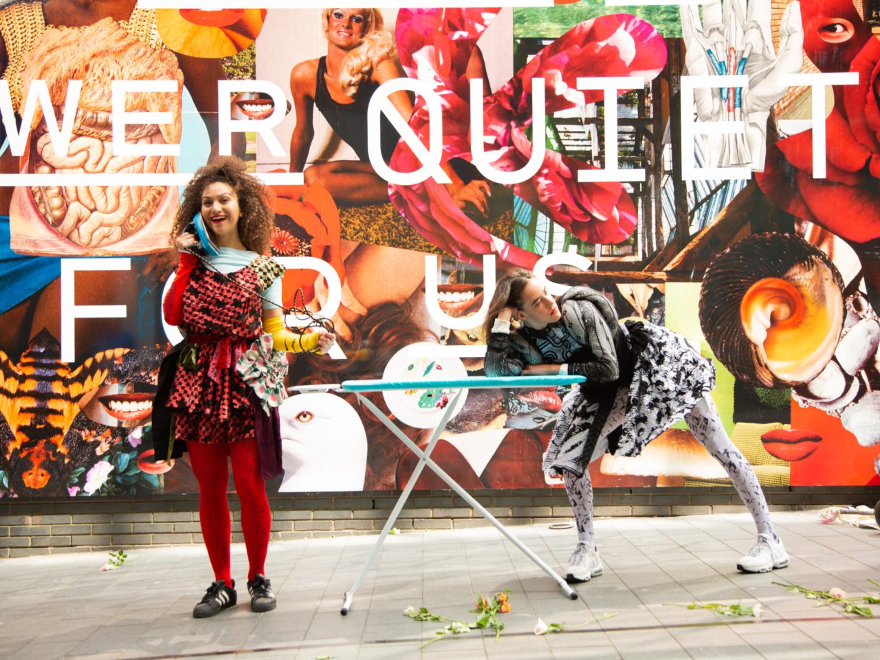 Two people performing, one person has an unplugged iron pressed up against their face as if they were on the phone and the other is leaning sideways on an ironing board.