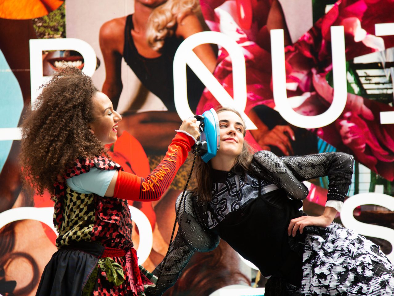 Two people performing in heavily patterned clothing, one person has an unplugged iron pressed up against the other persons face