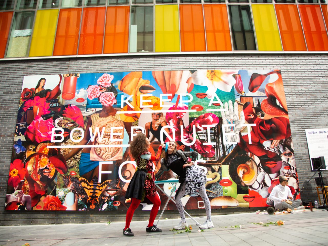 Two people performing with an iron and ironing board in front of a large wall collage.