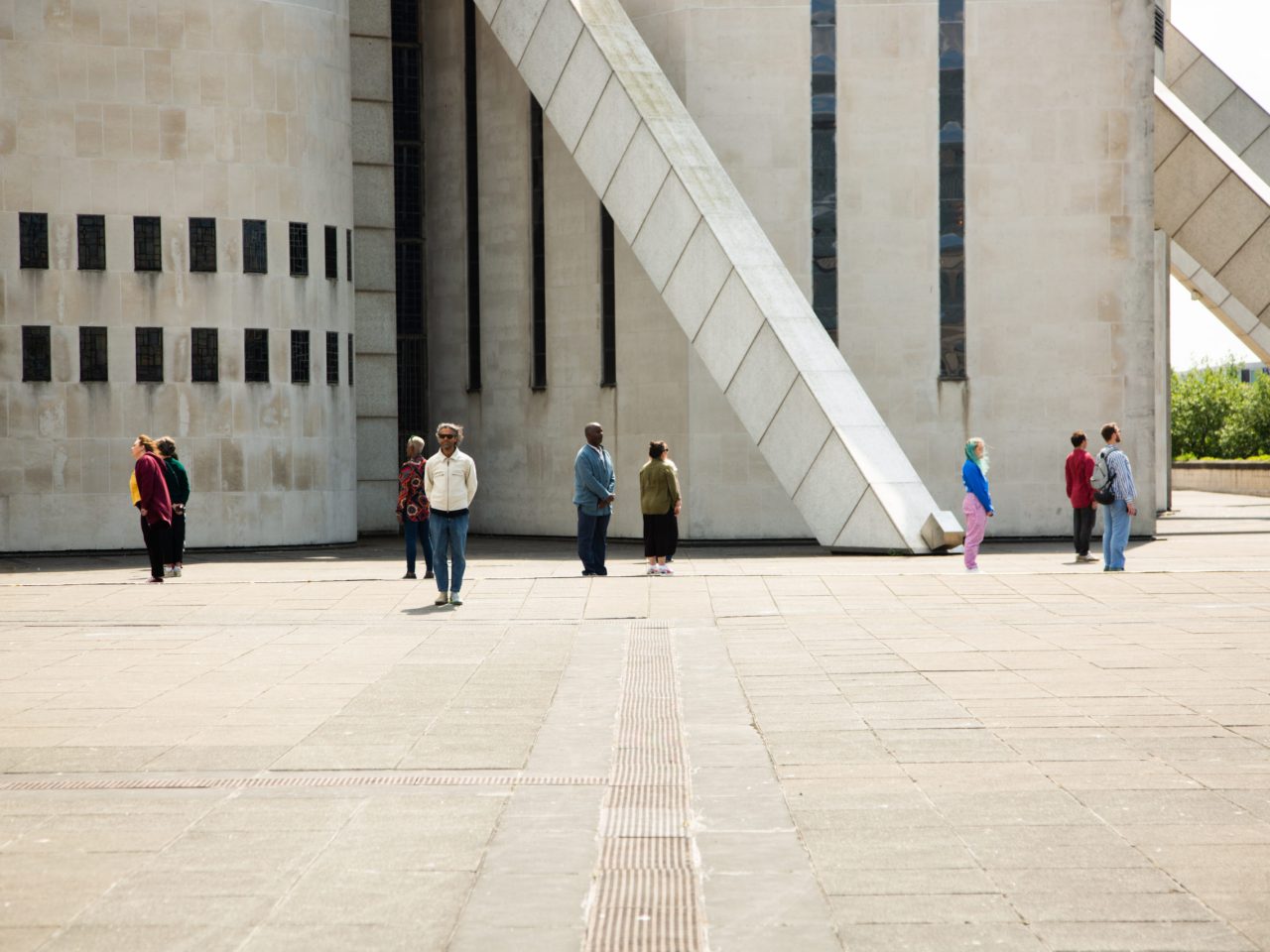 People standing equal distance apart, all turning away from each other. They are standing outside Liverpool Metropolitan Cathedral.