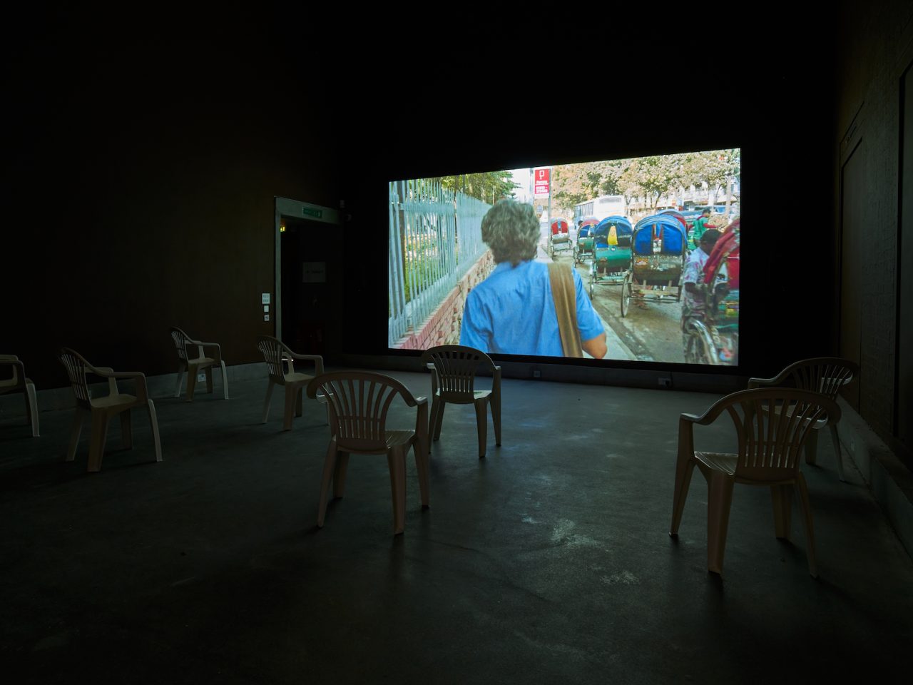 An installation shot of a film projected onto a wall, with white garden chairs in the foreground. The film depicts the back of someones head while they walk down the street.