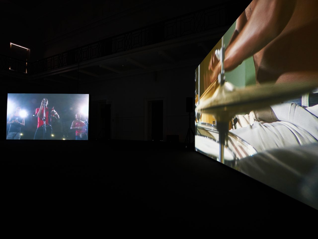 A two screen projection, one projection showing someone performing on stage with a mic and the other a close up of someone playing the drums.