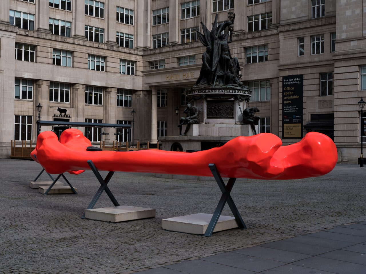 A installation within Exchange Flags, there is a bone shaped canoe, it is bright red and sat on a metal stand.
