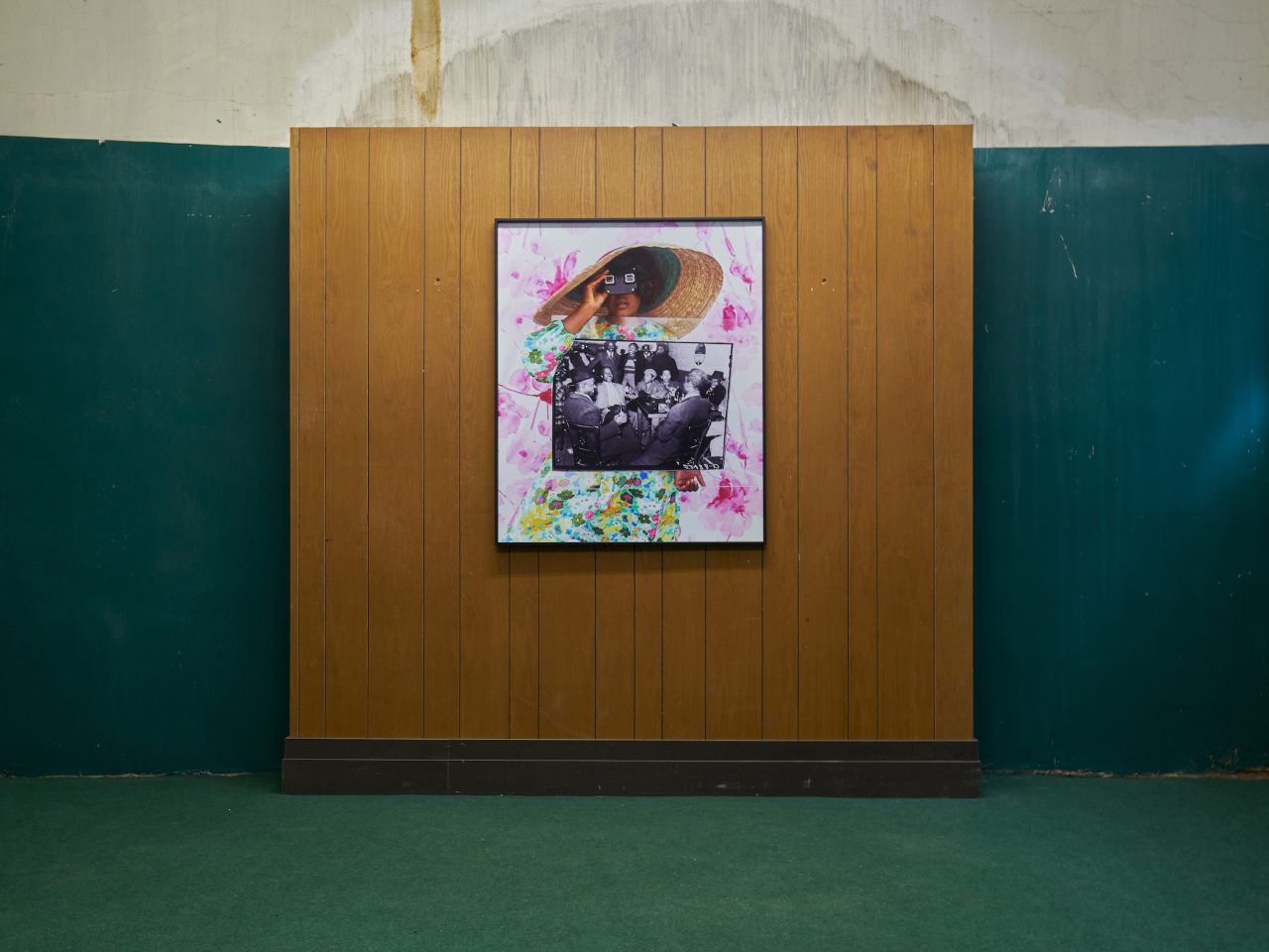 A framed artwork on the wooden wall. the wall is inbetween dark green walls and a green carpet. The artwork is a photo collage of an old black and white photograph amongst a flowery asset of a women looking towards the viewer.