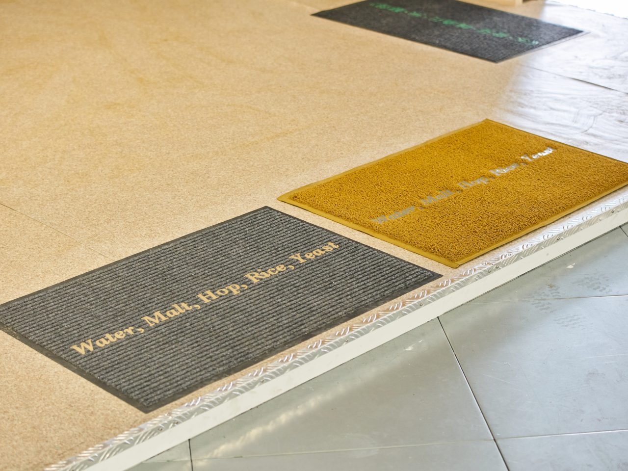 A grey and a yellow doormat, both reading 'Water, Malt, Hop, Rice, yeast'.