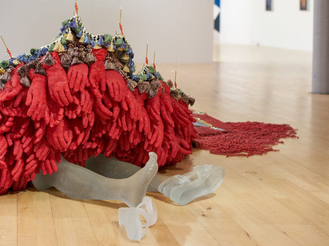 A sculpture of red gloves, lined with tassles. Coming from underneath are a persons legs lying down with their shoes coming off, the legs and shoes are made out of frosted glass.