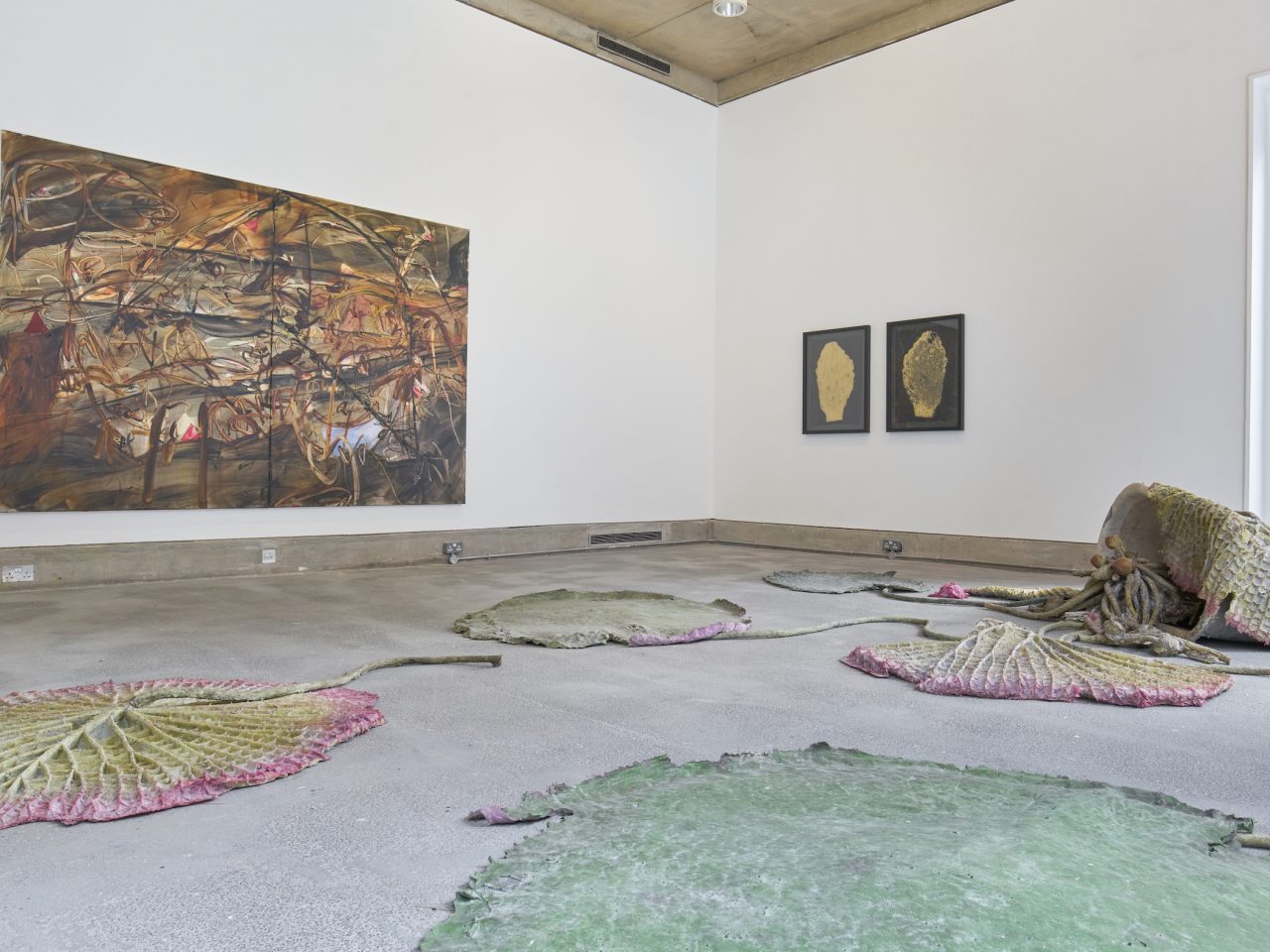 A large brown abstract painting on a white wall next to two framed prints of a gold leaf and there is a floor sculpture in the room.