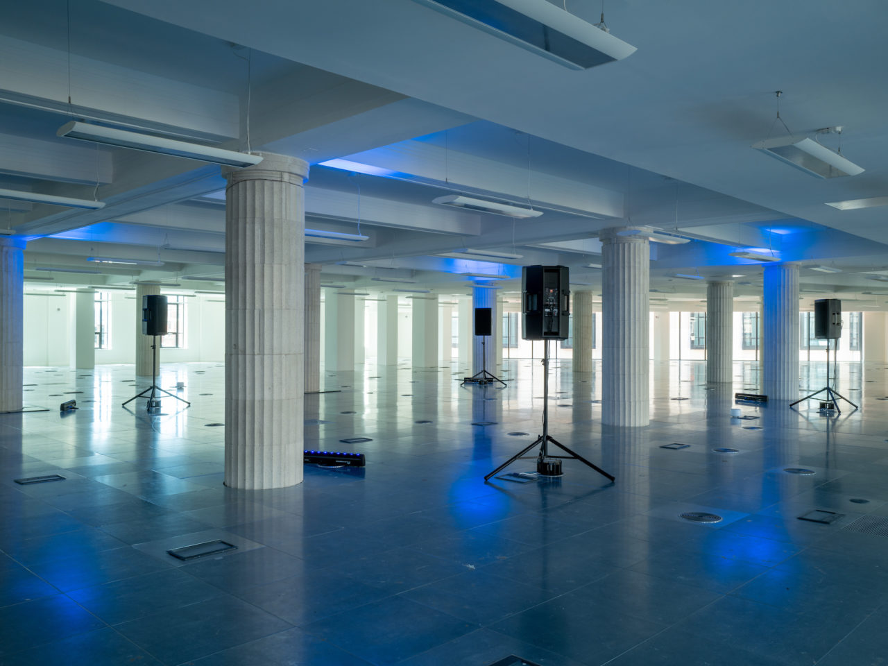 A large white room in the Lewis's Building with large white pillars throughout. In the middle of the room are speakers on stands all facing each other, the middle space is lit up with blue lighting and there is a sound piece playing.