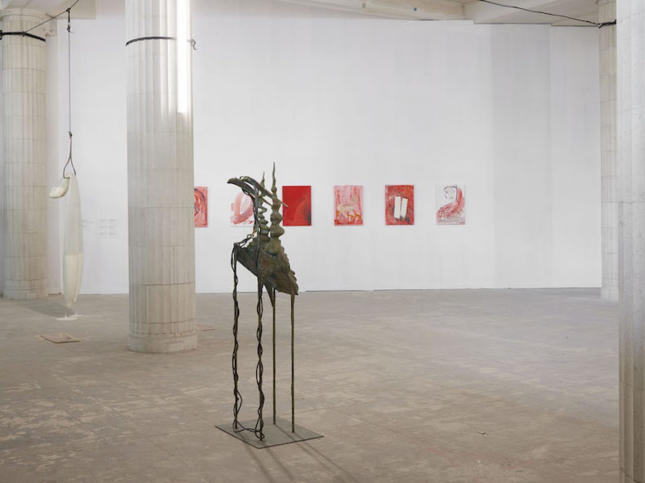 An abstract bronze sculpture in the middle of the space in front of a wall with small red abstract paintings