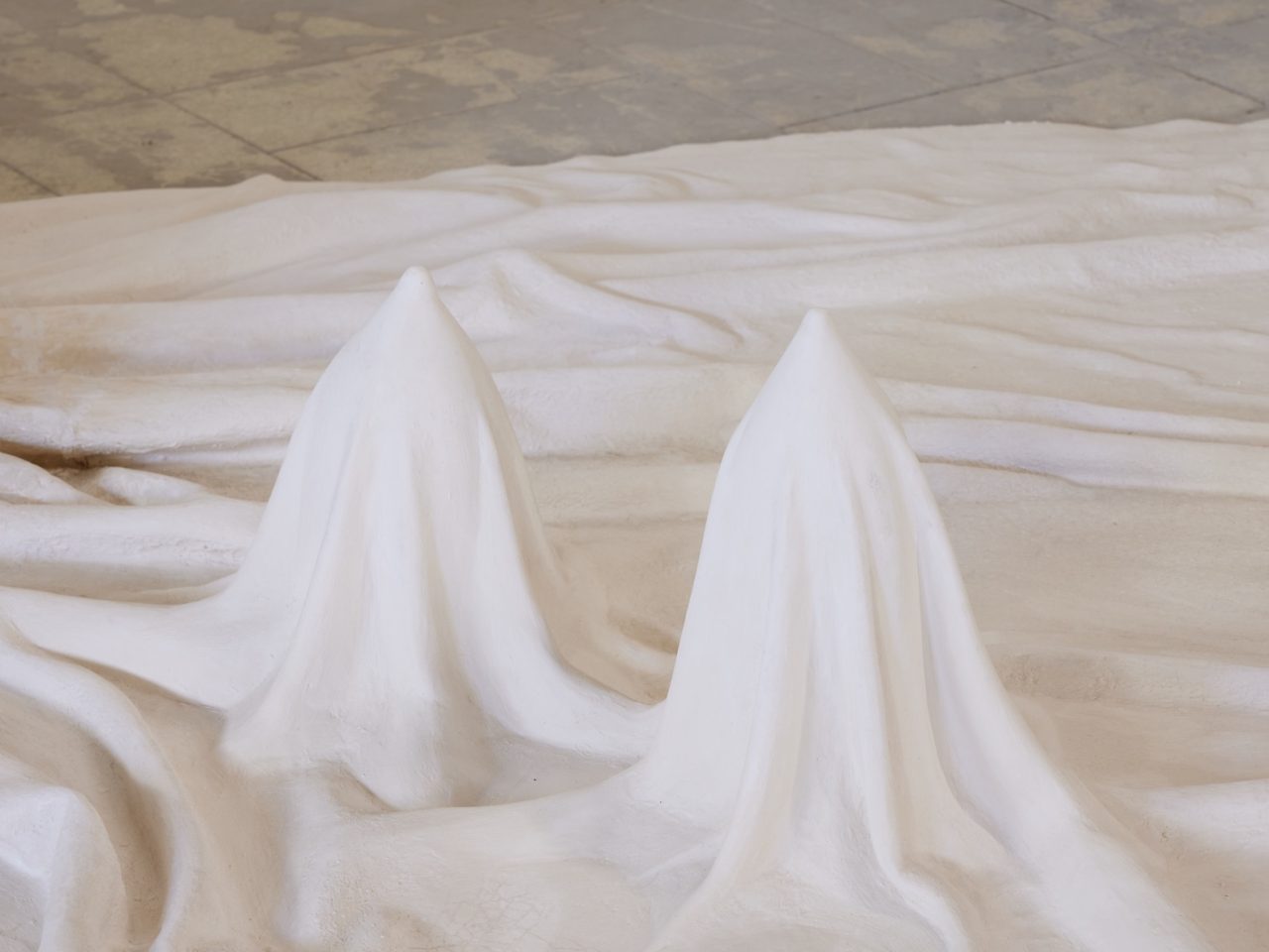 A white floor sculpture made to look like fabric draped across the floor. Within the sculpture there are two round and pointed shapes elevating as if they were coming from beneath the fabric.
