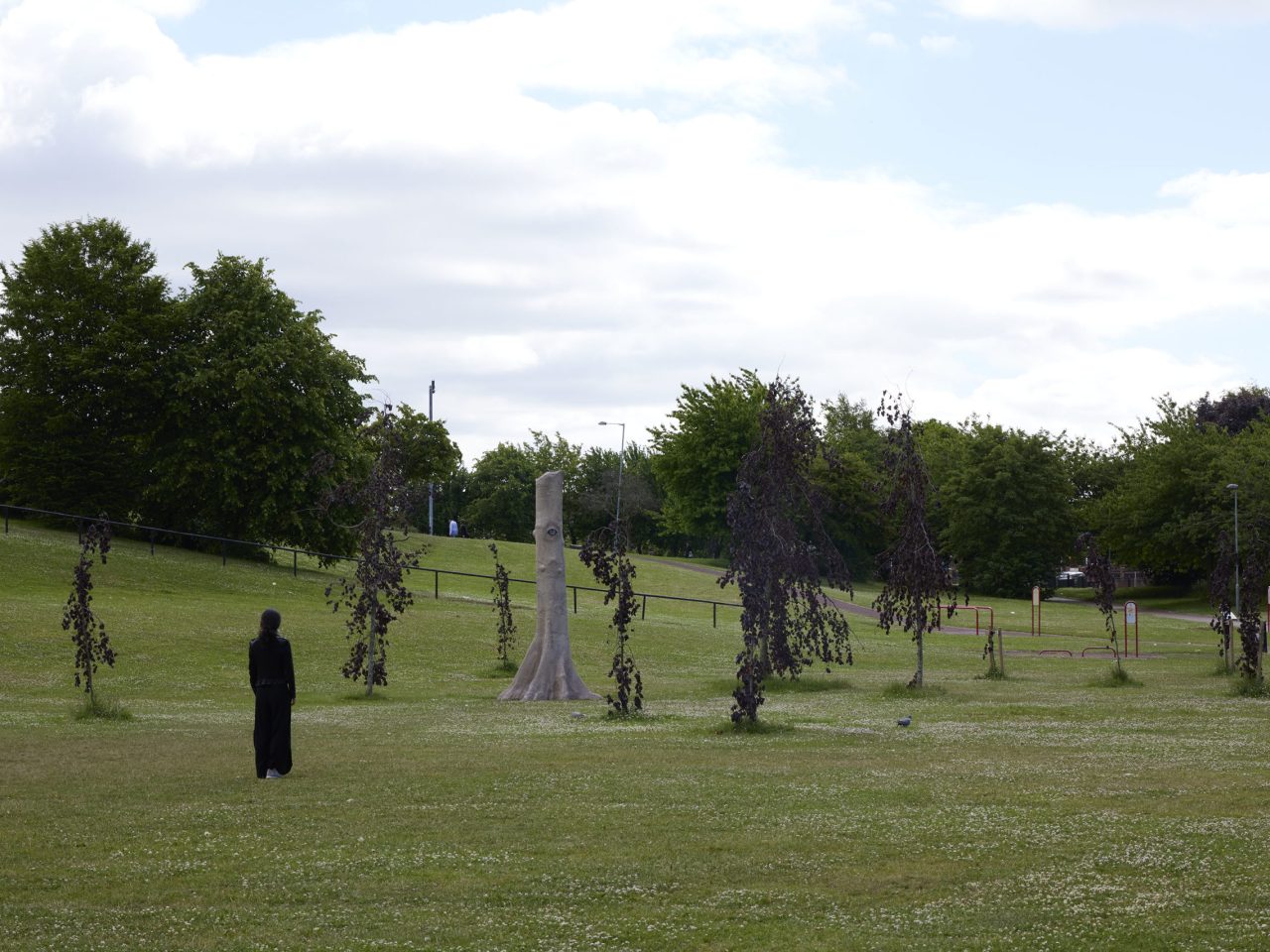 A sculpture amongst some trees on a feild, the sculpture is made to disguise as a tree and has a realistic orange/brown eye.