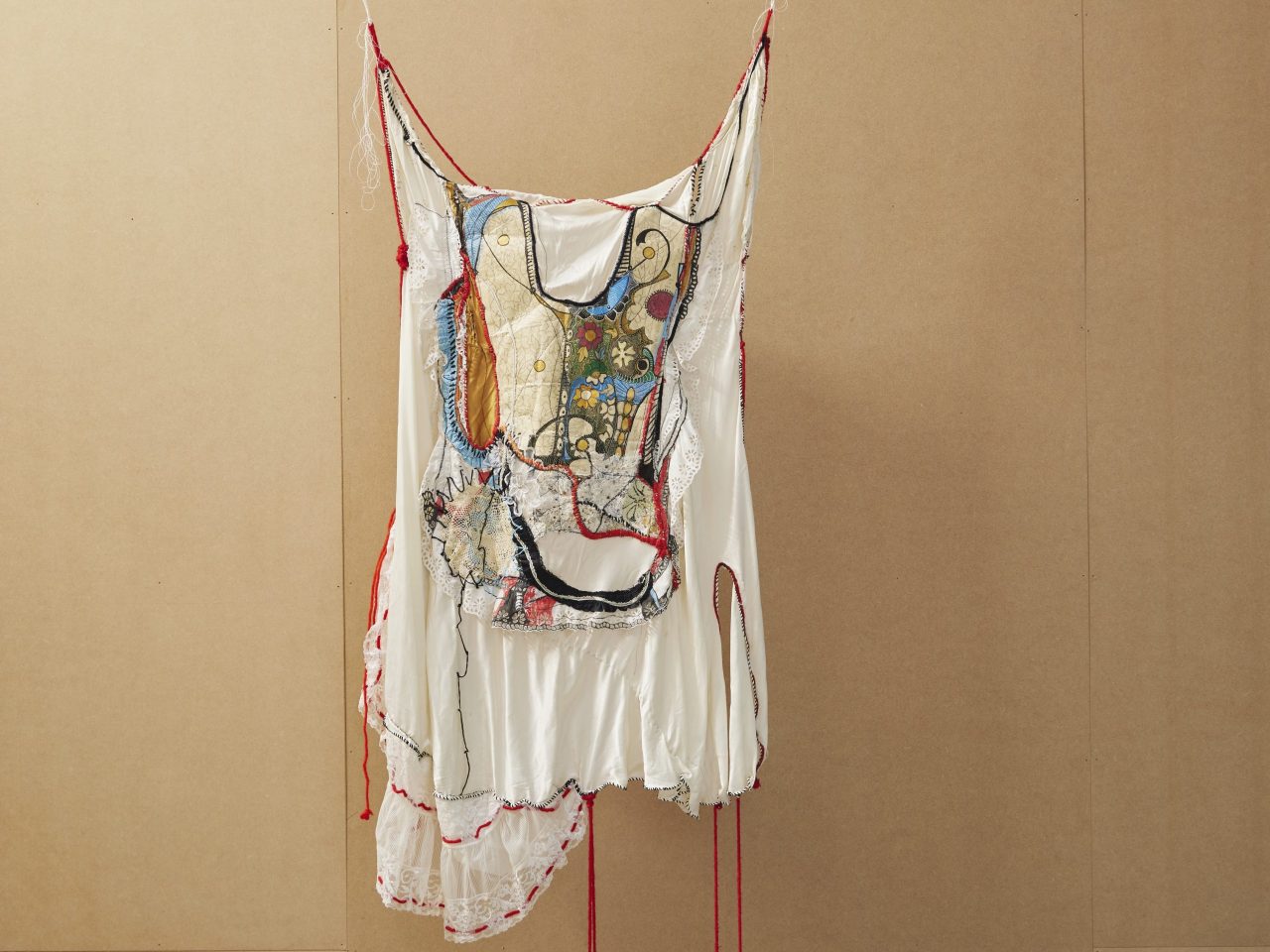 A hanging textile piece. It is white a draped white fabric with patterned fabric and stitching, and red threads hanging off the piece.