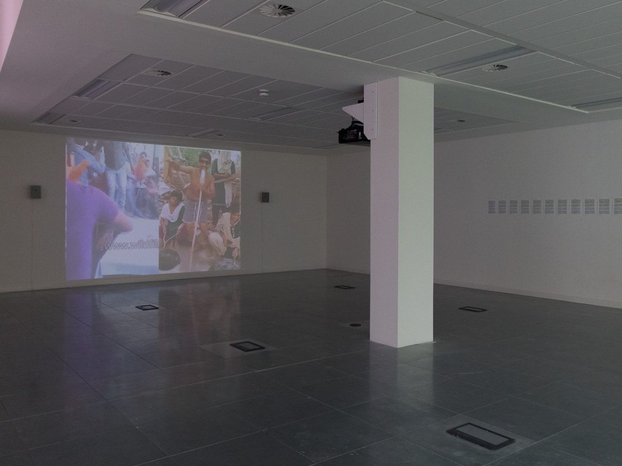 A large white room with a pillar in the centre and a projection on the wall of a spit screen showing a crowd of people.