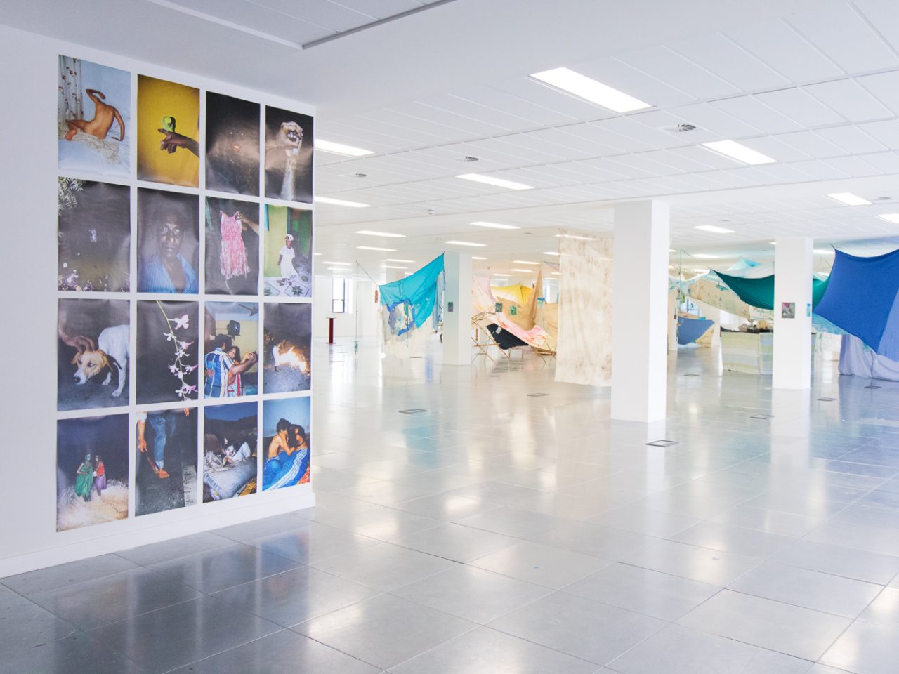 A large white room with an installation of fabrics suspended from the ceiling and a series of photographs mounted on the wall.
