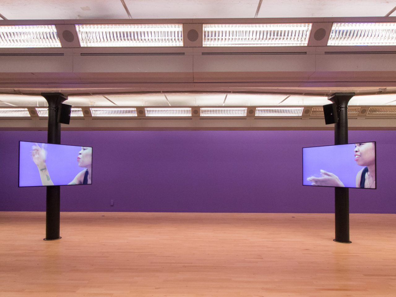 a gallery with purple walls and two screens. The screens show close up videos of the screens interacting with each other. The background of the videos, are the same purple as the gallery's.