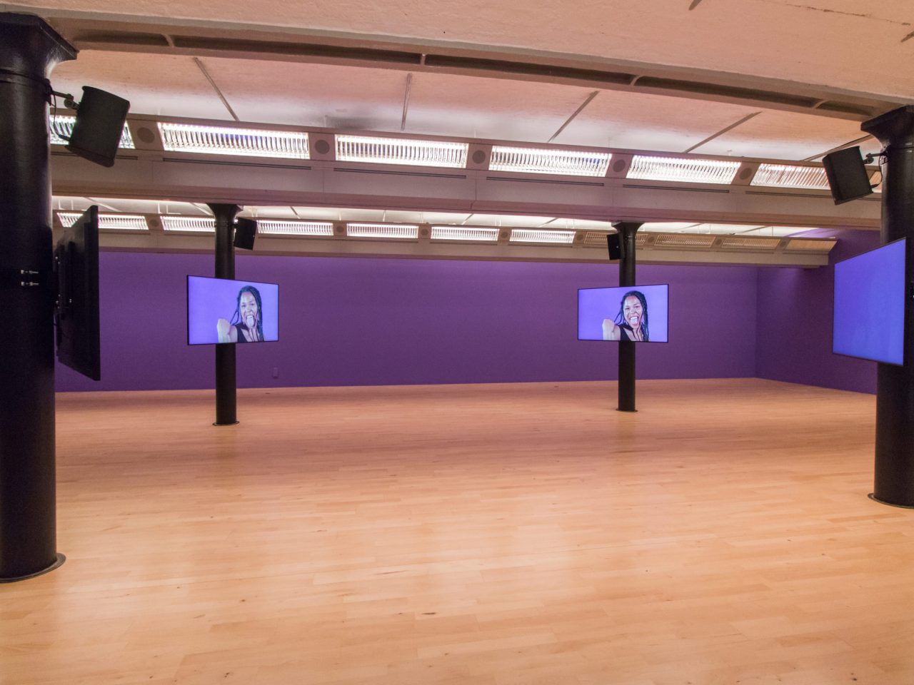 a gallery with purple walls and four screens. The screens show close up videos of the screens interacting with each other. The background of the videos, are the same purple as the gallery's.