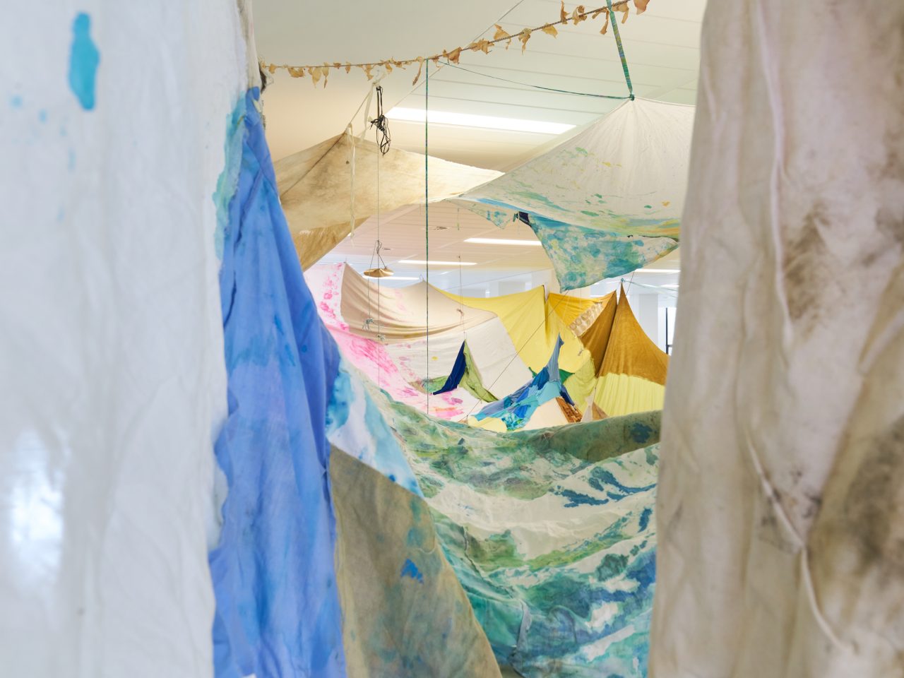 An large room installation of tie-dyed fabrics, stitched together as one piece, the piece covers the entire room and is hung irregularly to the ceiling.