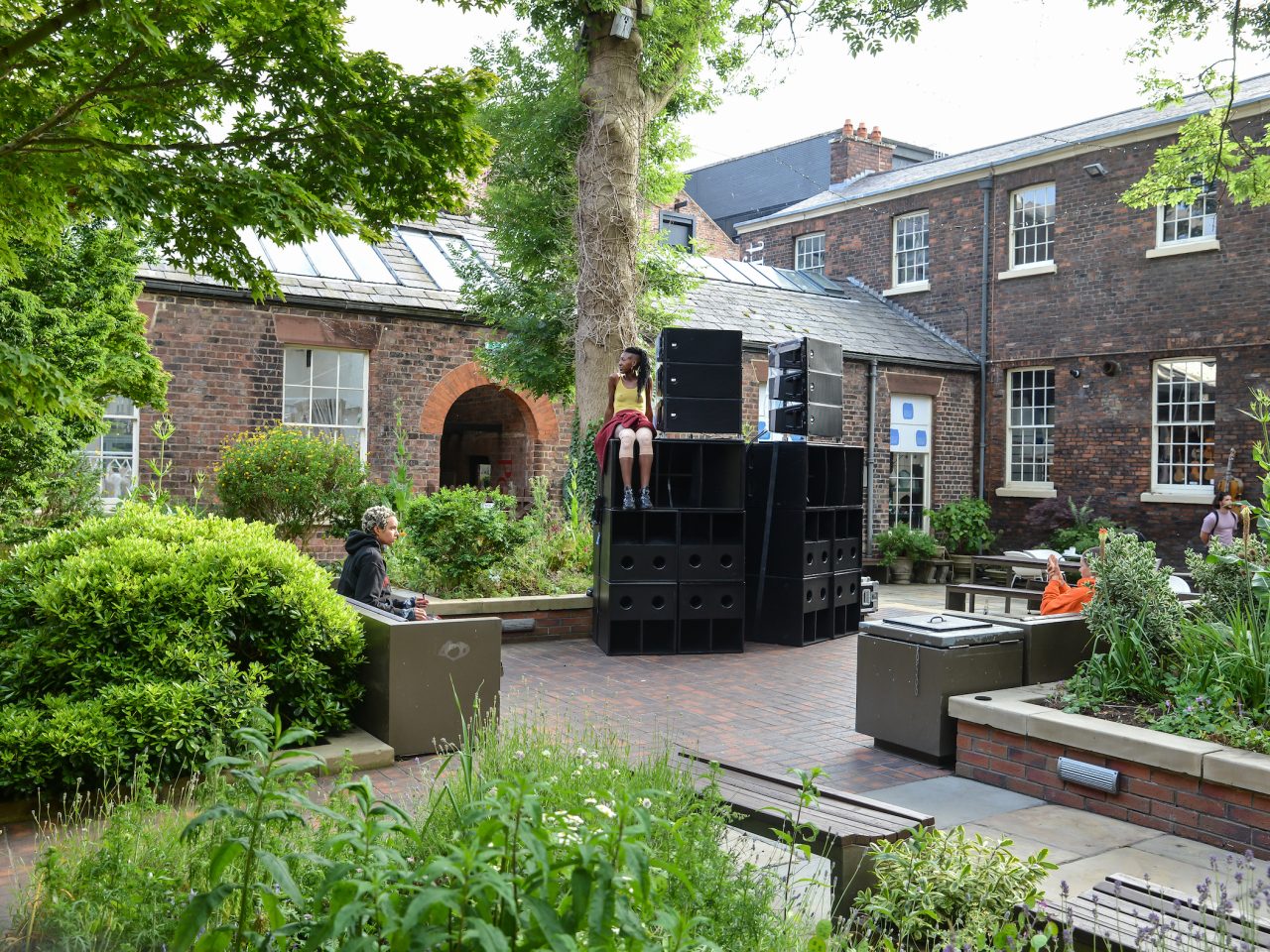 The artist sitting on a large speaker in a courtyard with people around watching.
