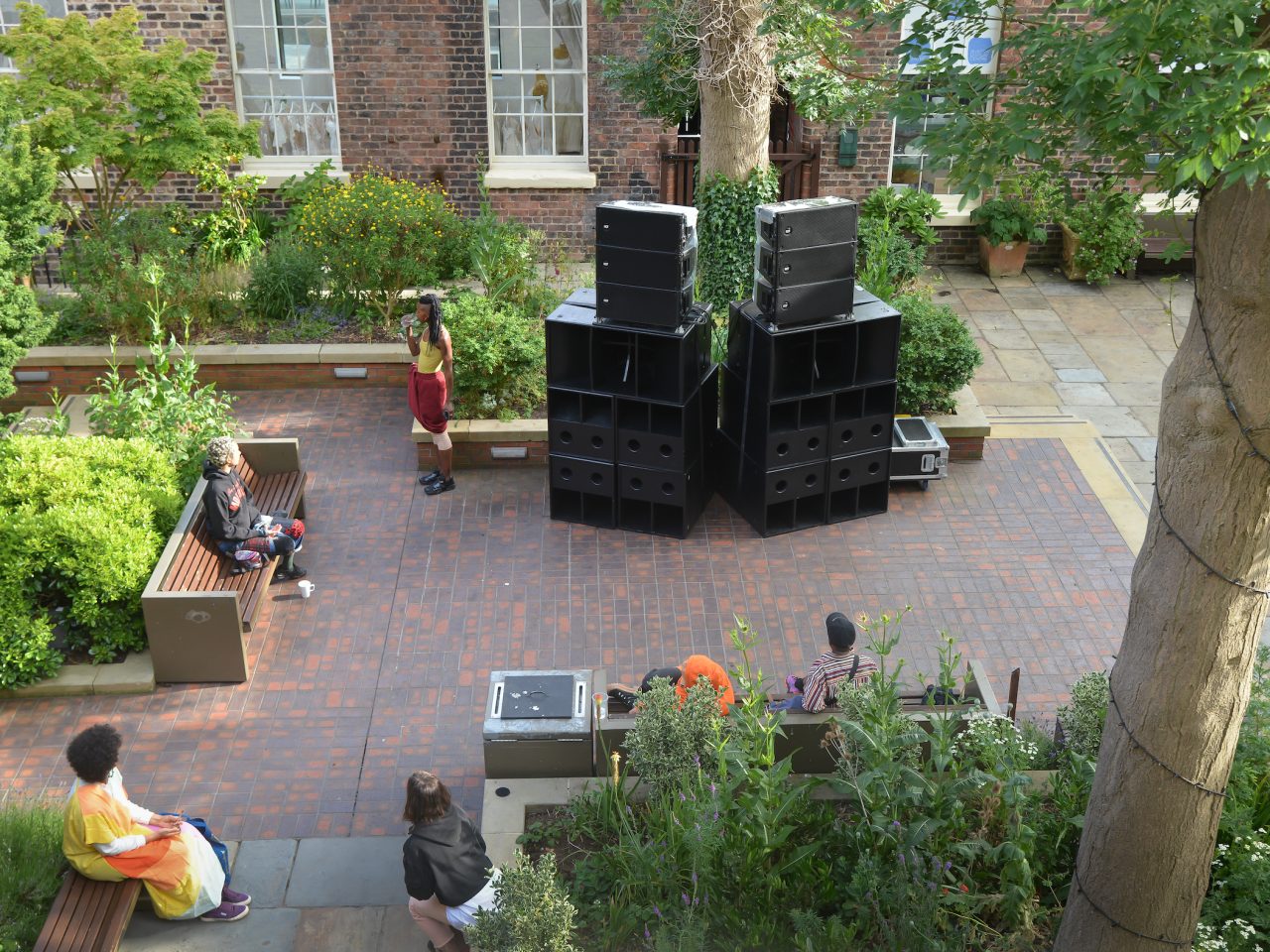 The artist speaking into a microphone, next to large speakers in a courtyard with people around watching.