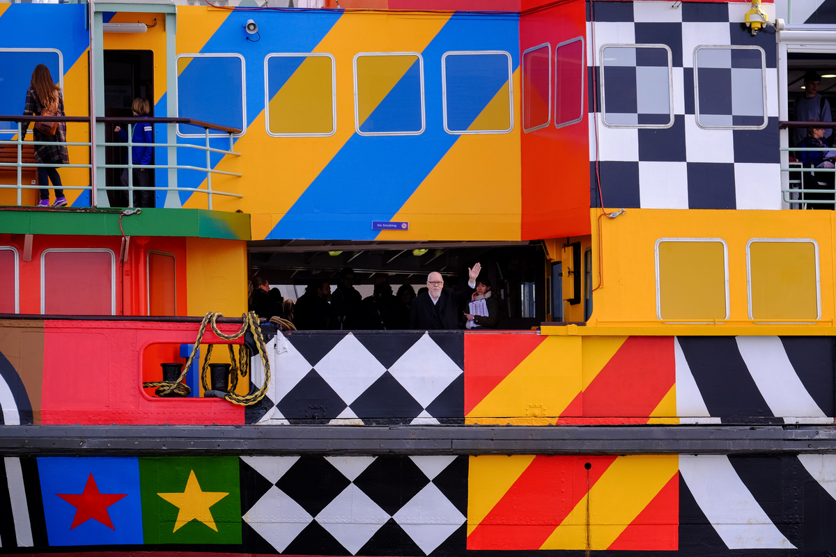 A close up shot of a colourful patterned ferry. The artist is on the ferry and waving to the camera