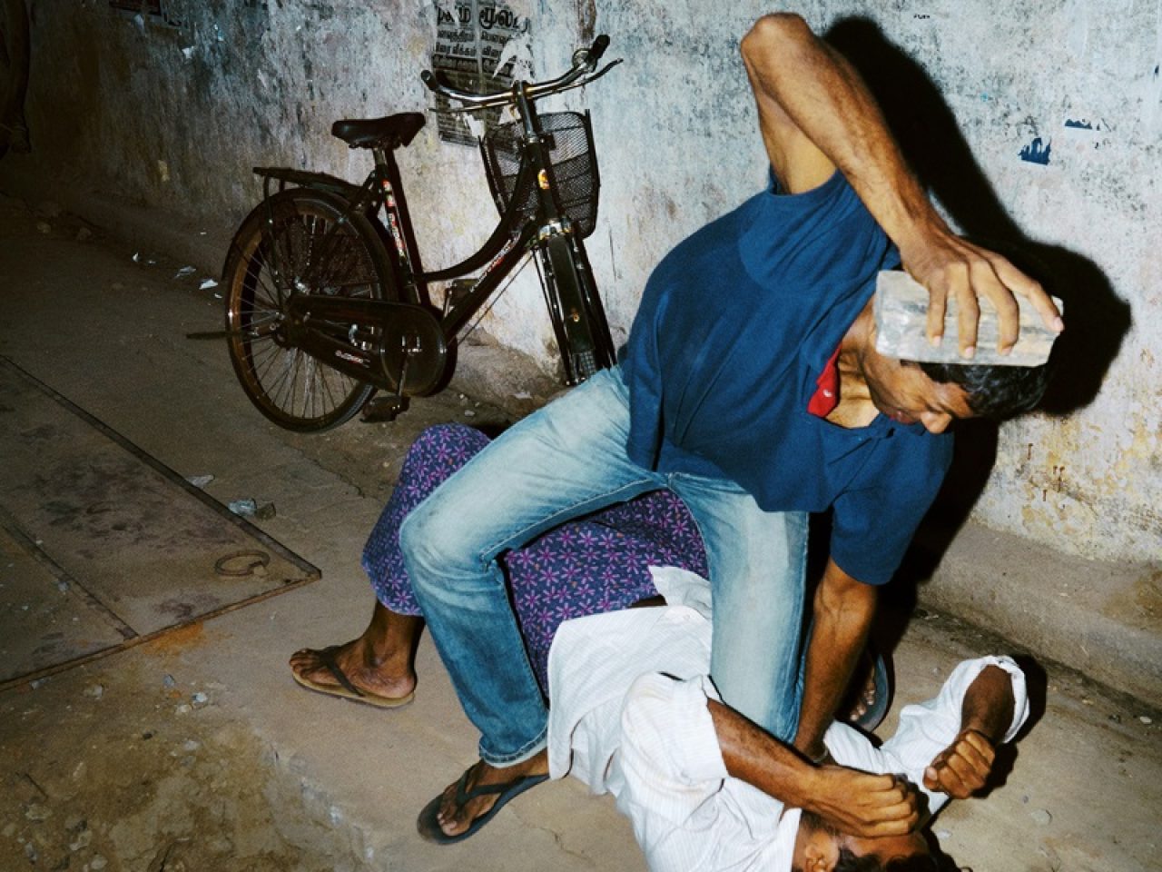 Two people fighting, one if on top of the other with a brick in their hand.