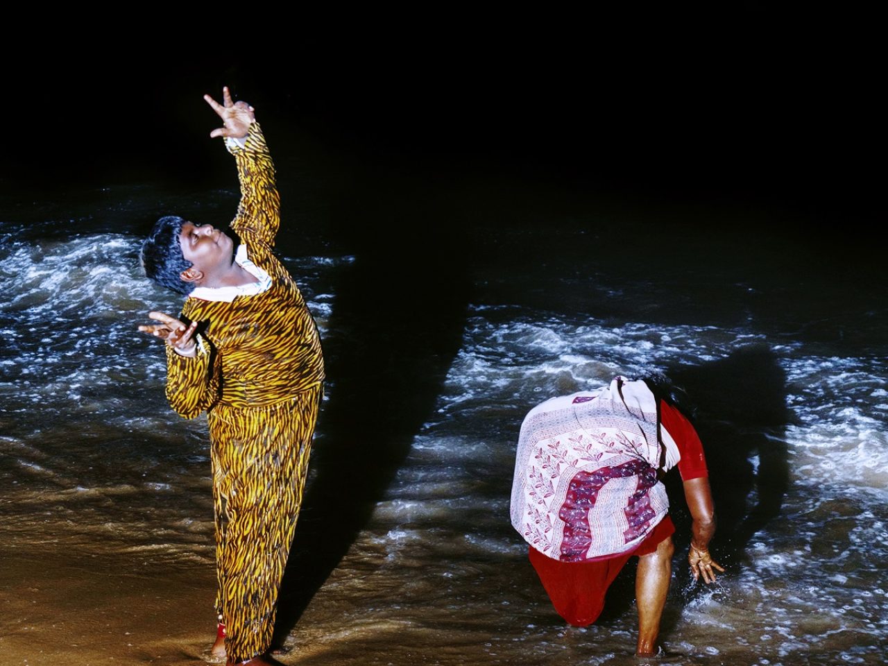 Two people on the coast of the beach, one is looking up with one hand up and the other is bending down.