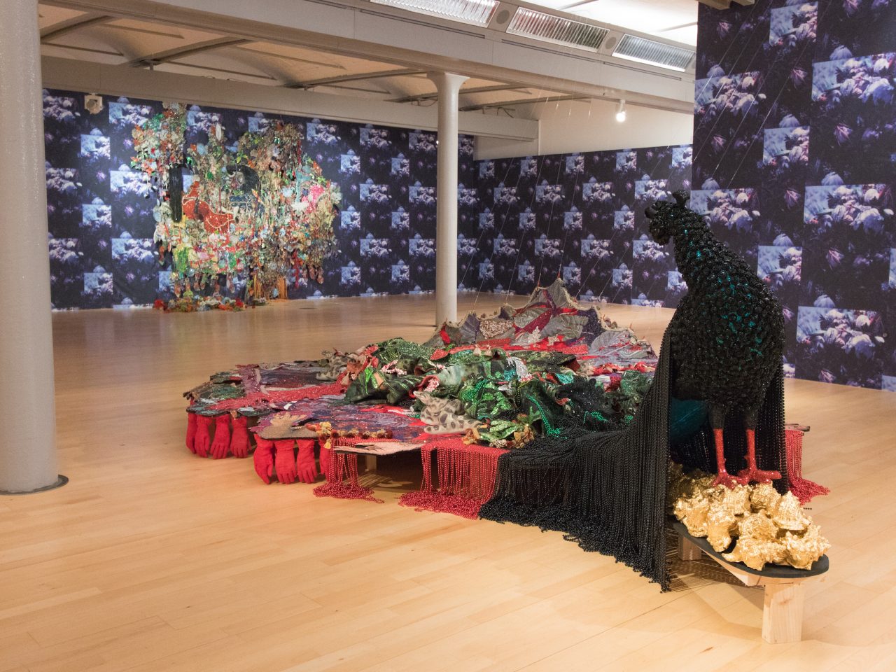 Sculpture of a preacock, the tail being a platform made up of purple, green and red fabric and beads. Underneath are stacked red gloves, lined with tassles. Coming from underneath the gloves are a persons legs lying down with their shoes coming off, the legs and shoes are made out of frosted glass.