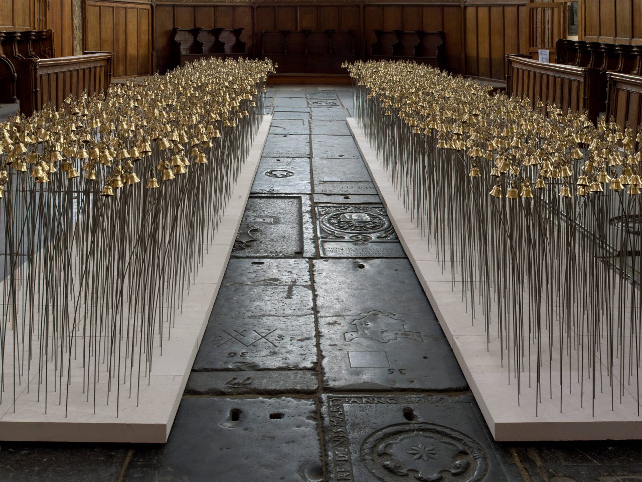 An installation of 2 long pathways of bells. It has a walkway in the middle so you can walk through it, this installation is inside a church⁠