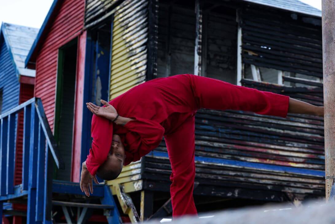A performer dancing, they have red overalls on. They are positioned similar to a capital T shape, with one leg lifted to the side and theire head and hands on the other side