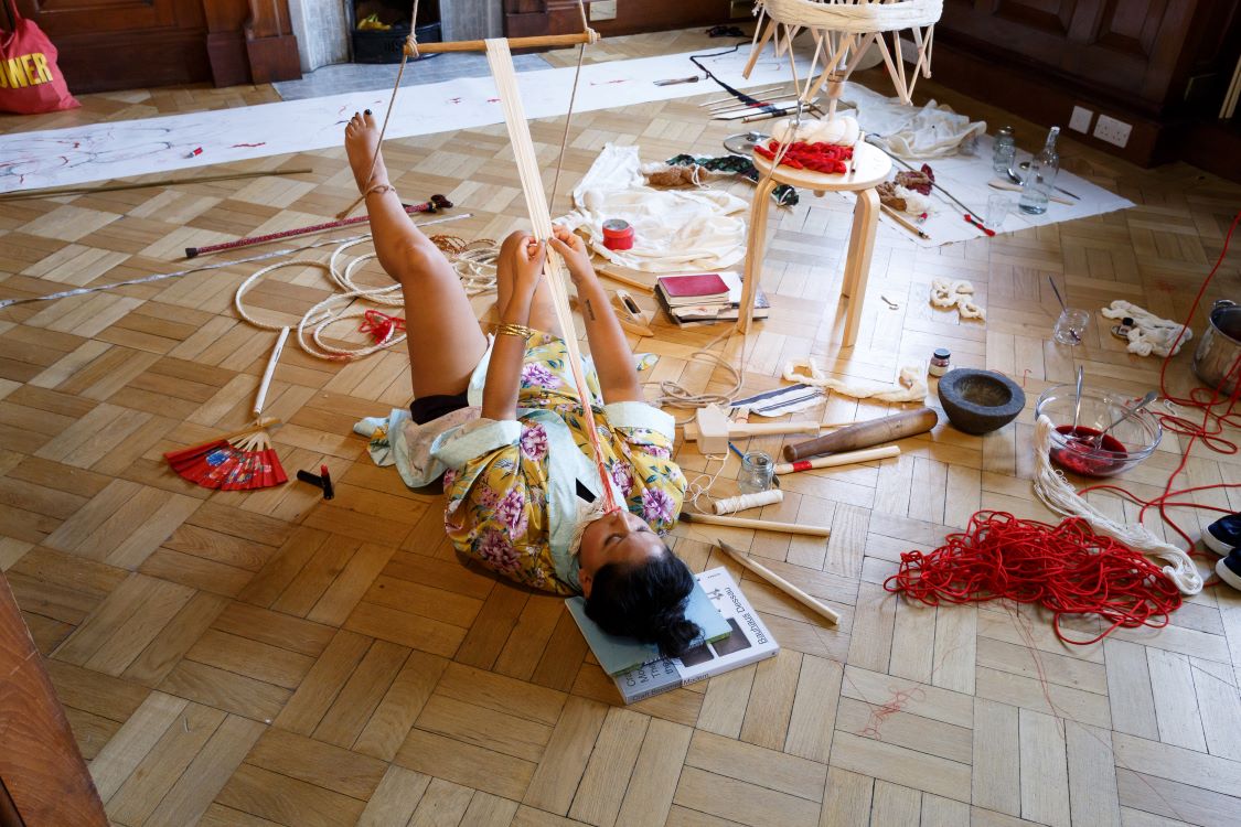 The artist is on their back with one leg held up by a rope; they are holding together a long thing piece of fabric in their mouth, which is connected to a wooden stick. Around them are many different materials, all items that are part of the artist's process.